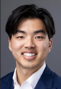 Smiling man in a blue suit with a white shirt, professional headshot on a gray background.