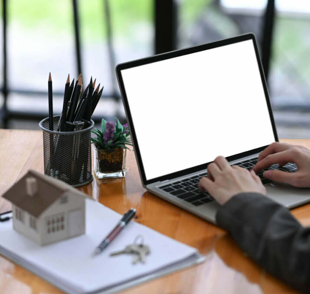 Real estate agent using laptop at wooden desk with house model and keys, enhancing property business efficiency.